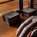 A close-up view of a tabletop scene featuring two small, wooden treasure chests with metal accents, each adorned with pirate-themed symbols. In the background, a tall, wooden structure resembling a ship's mast is partially visible, along with a human hand resting on the table, suggesting engagement with the game. The warm wooden surface adds to the overall pirate aesthetic of the setup.