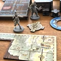 Two pirate miniatures are prominently displayed on a wooden table: one is a skeletal figure with a sword, and the other is a pirate with a tricorn hat and a hook for a hand. Nearby, a compass rose and a game board section titled "Time O'Movement" are visible, featuring a map with a ship illustration. In the background, there are wooden crates and a book labeled "DANGER - EXPLOSIVE," adding to the thematic setting of a pirate adventure.
