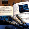A tabletop scene featuring a partially open rulebook for the game "Boarders and Black Flags," displaying sections on Skelliwags and Rapskullions, with accompanying illustrations of pirate miniatures. In the foreground, a skeletal hand is positioned, adding a thematic touch to the pirate aesthetic. Behind the book, several pirate miniatures are arranged, showcasing their detailed sculpts, including a mix of human and undead figures.