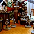 A collection of detailed miniatures from the game "Boarders and Black Flags" is displayed on a wooden surface, featuring both human pirates and undead Skelliwags. The miniatures are unpainted and vary in poses, with some wielding swords and others appearing to engage in action. In the background, a vibrant pirate-themed backdrop showcases a skull and crossbones design, along with elements of a ship, enhancing the thematic atmosphere of the game.