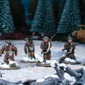 A group of six multi-part British and Canadian winter infantry figures stands on a snowy terrain, surrounded by evergreen trees and a tank in the background. Each figure is dressed in warm winter gear, showcasing a variety of poses and equipped with different weapons, including rifles and sidearms. The scene captures the essence of a winter battlefield, with patches of snow and scattered debris enhancing the atmosphere.