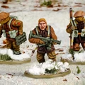 Three multi-part British and Canadian winter infantry figures are depicted in a snowy landscape. The central figure, with short blond hair, is holding a rifle and wearing a brown coat, while the other two figures are dressed in winter gear, one carrying a set of explosives and the other holding a sidearm. The bases of the figures are adorned with patches of snow and tufts of grass, enhancing the wintery scene.