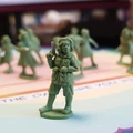 A detailed miniature figure of a woman stands prominently in the foreground, holding a pistol and wearing a military-style uniform, complete with a belt and pockets. Behind her, several other smaller green miniatures depict women in various poses, suggesting a scene of resistance. The background features a colorful tabletop surface with text, adding context to the setting.