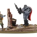 A detailed miniature scene features a soldier in military attire, holding a futuristic weapon, standing beside a crumbling brick wall. Opposite him, a heavily armored mech with a prominent red cape stands confidently, brandishing a spear-like weapon. The base is textured with grass and rocks, enhancing the dynamic and gritty atmosphere of the setting.