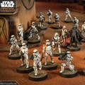 A group of Star Wars miniatures features several Stormtroopers in white armor, positioned in a desert-like environment with a backdrop of a weathered building. Among them stands a prominent figure in black armor, holding a red lightsaber, likely representing Darth Vader. The scene is detailed with various poses of the troopers, some aiming weapons, while others appear to be in action, all set on circular bases with textured surfaces.