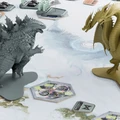 A detailed tabletop scene features two large miniatures: Godzilla in a gray color and King Ghidorah in a tan color, positioned on a game board with various hexagonal tiles. Surrounding them are additional game elements, including armored vehicles and other tokens, set against a backdrop that suggests a dynamic battlefield. The overall composition highlights the cinematic combat theme of the game, emphasizing the iconic nature of the monsters.