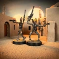 Two IG-Series Assassin Droids stand in a sandy, desert-like environment, each on a round black base. The droids are metallic with intricate detailing, one holding a blaster in each hand, while the other raises a weapon high. The background features a stylized, sci-fi setting with structures and soft lighting, evoking a Star Wars atmosphere.