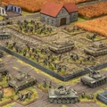A detailed tabletop wargaming scene depicts a battlefield layout featuring American and German forces from the Flames Of War starter set, Fortress Europe. In the foreground, several M4 Sherman tanks and infantry figures are positioned, while in the background, a mix of German Panzergrenadiers and tanks, including Tiger tanks, are arranged near a small building with a red roof. The terrain includes fields, roads, and scattered structures, creating a dynamic and immersive environment for gameplay.