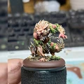 A detailed miniature of a fierce creature stands on a circular base, showcasing a blend of organic and monstrous features. The creature has a muscular build, covered in green foliage and sharp spikes, with a wild mane of hair and glowing red eyes that convey a menacing presence. Its pose suggests an aggressive stance, ready to pounce, with a background of blurred keyboard keys, indicating a workspace setting.