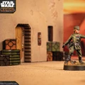 A detailed miniature of a customizable Imperial Officer stands prominently in the foreground, wearing a green uniform with a rank insignia and a cap. The background features a sandy terrain with crates and a building entrance, suggesting a desert environment typical of the Star Wars universe. The scene is set within a tabletop gaming context, enhancing the immersive experience for players.