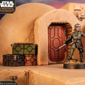 A detailed miniature of a character stands in front of a rounded, tan structure resembling a building from the Star Wars universe, with a red door and various mechanical elements visible. The character is dressed in a green uniform, holding a weapon, and is positioned on a rocky base. Nearby, there are several colorful crates in green, orange, and black, adding to the scene's immersive atmosphere.
