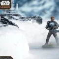A detailed miniature of a character from the Star Wars universe stands prominently in a snowy landscape, holding a weapon with a focused expression. The character has distinctive features, including a unique head shape and is dressed in a blue and brown outfit with armor. In the background, blurred outlines of rocky terrain and a lantern can be seen, enhancing the atmospheric setting.