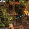 A detailed miniature of Luke Skywalker stands on a rocky terrain, holding a blue lightsaber in one hand and a blaster in the other. He is dressed in a yellow jacket with black accents, and the background features lush greenery and wooden structures, creating a vibrant and dynamic scene. The image captures the essence of the Star Wars universe, highlighting the character's iconic look and the adventurous setting.