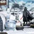 A detailed scene from Star Wars Legion features iconic miniatures set in a snowy landscape. Prominently displayed are Darth Vader, holding a red lightsaber, and a uniformed officer pointing, surrounded by several Stormtroopers. The terrain includes patches of grass and rocky elements, enhancing the immersive atmosphere of the battlefield.