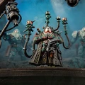 A detailed miniature figure stands prominently in the foreground, featuring a character with a complex helmet and multiple mechanical appendages extending from its back, each topped with small, glowing orbs. The figure is dressed in a dark, ornate robe, holding a device in one hand, while two other armored figures are partially visible in the background, suggesting a sci-fi setting. The scene is set against a moody, atmospheric backdrop that enhances the futuristic theme.