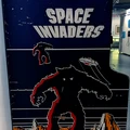 A vintage arcade cabinet for the game "Space Invaders" features a striking design with bold, white lettering at the top. The artwork depicts a large, menacing alien creature in black and red, with smaller figures and a spaceship above it, set against a dark blue background. The cabinet shows signs of wear, adding to its nostalgic charm, and is positioned in a well-lit space that suggests a gaming or exhibition environment.