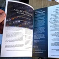 A hand holds open a magazine titled "Secret Passages," revealing two pages. The left page features an advertisement for "Tabletop Sentinel," promoting daily gaming and industry news, with a colorful collage of tabletop gaming images in the background. The right page introduces "Secret Passages," outlining its content and welcoming readers, with a list of articles and topics covered, including references to various tabletop games and cultural commentary. The overall design is clean and visually engaging, with a mix of text and imagery.