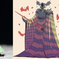 A green resin miniature of a character stands on the left, showcasing intricate details and a dynamic pose. On the right, a colorful illustration depicts the same character, featuring a patterned cloak in shades of purple and green, a helmet with bat-like features, and a menacing expression. The character wields a staff with a red tip, surrounded by splashes of red, enhancing the dramatic effect.