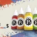 Four bottles of P3 paint are displayed against a colorful, textured background. The bottles include "Gunner Red," "Menite Ochre," "Radiant Yellow," and "Witchfire Green," each featuring a black gear logo with "P3" prominently displayed. The background features splashes of blue, red, and white paint, adding a vibrant artistic flair to the scene.