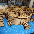 A detailed model of the TTCombat Mega Turbine, featuring a circular base with multiple levels and intricate cut-out patterns. The structure includes staircases leading up to the main platform, which is adorned with various mechanical details and a central turbine section. The model is set against a blue cutting mat, with tools and paint supplies visible in the background, indicating an active workspace.