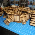 A detailed assembly of the TTCombat Mega Turbine showcases its intricate design, featuring layered MDF components with a mix of circular and rectangular shapes. The structure includes multiple staircases leading up to a central platform, with various pipes and supports adding to its industrial aesthetic. The backdrop consists of a blue cutting mat, indicating the workspace, and shelves filled with paint bottles, suggesting an active modeling environment.
