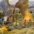 A detailed diorama depicts a World War I battlefield scene with soldiers in blue uniforms engaged in combat amidst smoke and explosions. In the background, a landscape of rolling hills is visible, with barbed wire and trees partially obscured by billowing smoke. The foreground features vibrant yellow foliage, enhancing the dramatic atmosphere of the scene.
