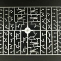 A plastic sprue featuring various components for miniature figures, including multiple torsos, limbs, and heads designed for 28mm scale modern civilians and post-apocalyptic survivors. The figures are in a gray plastic, arranged in a grid pattern, showcasing a variety of poses and accessories. The central part of the sprue includes a circular connector, with the pieces surrounding it neatly organized for easy access during assembly.