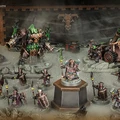 A detailed display of the new Zharrdron army features various miniatures set against a dark, industrial backdrop. In the center, a prominent War Despot stands on a pedestal, clad in imposing armor and wielding a staff, surrounded by Infernal Cohorts holding spears. To the left, a large, menacing Dominator Engine looms, while a mortar-like Tormentor Bombard is positioned on the right, showcasing the army's dark and chaotic aesthetic.