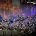 A dynamic scene features a group of intricately detailed Warhammer 40,000 miniatures engaged in battle. In the foreground, several armored soldiers in green uniforms are positioned behind a barricade, aiming their weapons. In the background, imposing figures in dark blue armor, adorned with spikes and intricate designs, advance menacingly, showcasing a mix of futuristic and gothic architecture in the blurred backdrop.