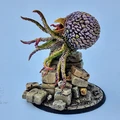 A detailed miniature of a creature resembling a large, bulbous head with a textured surface, featuring vibrant colors like purple and green. It has multiple long, tentacle-like appendages extending from its base, which is perched atop a pile of irregularly shaped stone blocks. The base is circular and textured, resembling a rocky terrain, enhancing the overall fantasy aesthetic of the piece.
