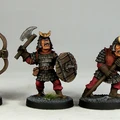 Three fantasy miniatures are displayed, each depicting a character in armor with distinct features. The first figure on the left holds a bow, while the middle figure wields an axe and a shield, showcasing a fierce expression. The third figure on the right is armed with a spear, all standing on textured bases with a hint of grass. Each miniature is painted with vibrant colors, emphasizing their detailed armor and facial expressions.