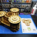 A detailed model of the Sector 3 Silo Hub from TT Combat is prominently displayed on a blue cutting mat. The structure features multiple cylindrical silos with intricate wooden textures and circular tops, showcasing a combination of open latticework and solid panels. In the background, various paint pots and a few other miniature models are visible, along with the instruction sheet for the Silo Hub, which includes an illustration of the assembled model.