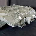 A large, detailed model of a spaceship, featuring a flat, ramp-like structure at the front and a complex array of panels and textures across its surface. The ship has a robust, angular design with various protrusions and openings, suggesting functionality for cargo or troop deployment. The model is presented in a light gray color against a dark background, highlighting its intricate details.