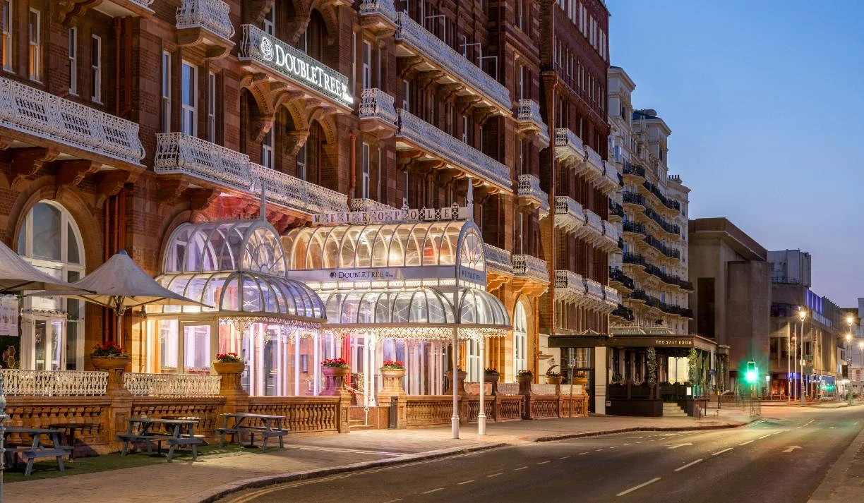 A grand hotel entrance features an ornate glass canopy with decorative lighting, flanked by brickwork and balconies of the building. The DoubleTree Metropole Hotel is prominently displayed above the entrance, while flower pots add a touch of color to the scene. The street is quiet, illuminated by soft evening lights, creating a serene atmosphere as dusk settles.