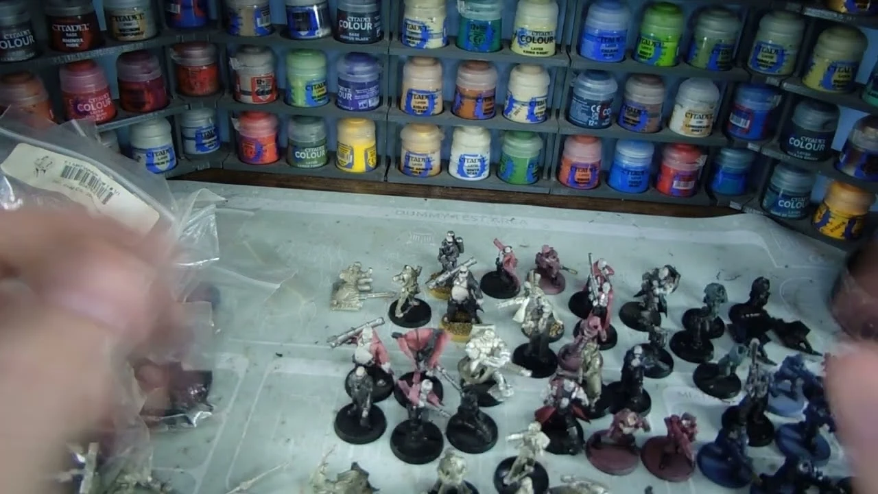 A variety of Heresy Miniatures models are arranged on a work surface, showcasing a mix of painted and unpainted figures in various poses. In the background, shelves filled with paint pots in a range of colors, including blues, greens, and reds, provide a vibrant backdrop. The models include soldiers and fantastical characters, some with detailed bases, while the surface beneath them is marked with paint splatters and tools of the hobby.