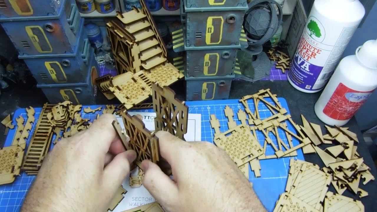A workspace filled with various pieces of laser-cut terrain and building components, primarily made of light brown wood. In the foreground, a hand is assembling parts, including a set of stairs and wall sections, while a blue cutting mat with grid lines provides a base. In the background, several painted structures are stacked, alongside bottles of glue and other crafting supplies.