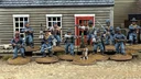A group of ten detailed 28mm scale miniatures representing dismounted US Cavalry and Buffalo Soldiers stands in front of a wooden building with a red door. The miniatures are posed with various weapons, including rifles and a bugle, showcasing a mix of uniforms and accessories, such as scarves and hats. A small beagle and a cactus are also present among the figures, all mounted on 25mm round bases with a sandy terrain effect.