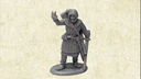 A grey resin miniature of the Master Fletcher stands on a circular base, depicted in a friendly pose with one hand raised in a gesture. He is dressed in a hooded cloak and tunic, with a quiver of arrows slung over his shoulder and a sword at his side. The background features a textured, light-colored surface that enhances the focus on the figure.