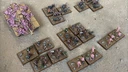A collection of Zayken miniatures from Void Scar Miniatures is displayed on a textured surface. In the foreground, several bases feature hordes of zombified figures in various colors, including green, purple, and pink, representing the Neophyte Mobs. To the left, a tank is adorned with grotesque, bloated creatures, suggesting a fusion of machine and undead, while additional bases of zombies are scattered throughout the scene, emphasizing the overwhelming numbers of the Zayken forces.