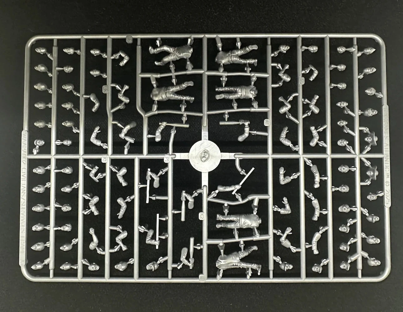 A plastic sprue featuring various components for miniature figures, including multiple torsos, limbs, and heads designed for 28mm scale modern civilians and post-apocalyptic survivors. The figures are in a gray plastic, arranged in a grid pattern, showcasing a variety of poses and accessories. The central part of the sprue includes a circular connector, with the pieces surrounding it neatly organized for easy access during assembly.