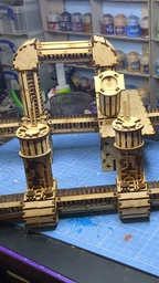 A detailed assembly of the TT Combat Sector 4 Plasma Relay is displayed, featuring intricate wooden structures with multiple towers and connecting pathways. The design includes cylindrical towers with circular tops and various platforms, all set against a blue cutting mat. In the background, jars of paint and tools are visible, indicating a workspace dedicated to tabletop gaming and model building.