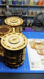 A detailed model of the Sector 3 Silo Hub from TT Combat is prominently displayed on a blue cutting mat. The structure features multiple cylindrical silos with intricate wooden textures and circular tops, showcasing a combination of open latticework and solid panels. In the background, various paint pots and a few other miniature models are visible, along with the instruction sheet for the Silo Hub, which includes an illustration of the assembled model.