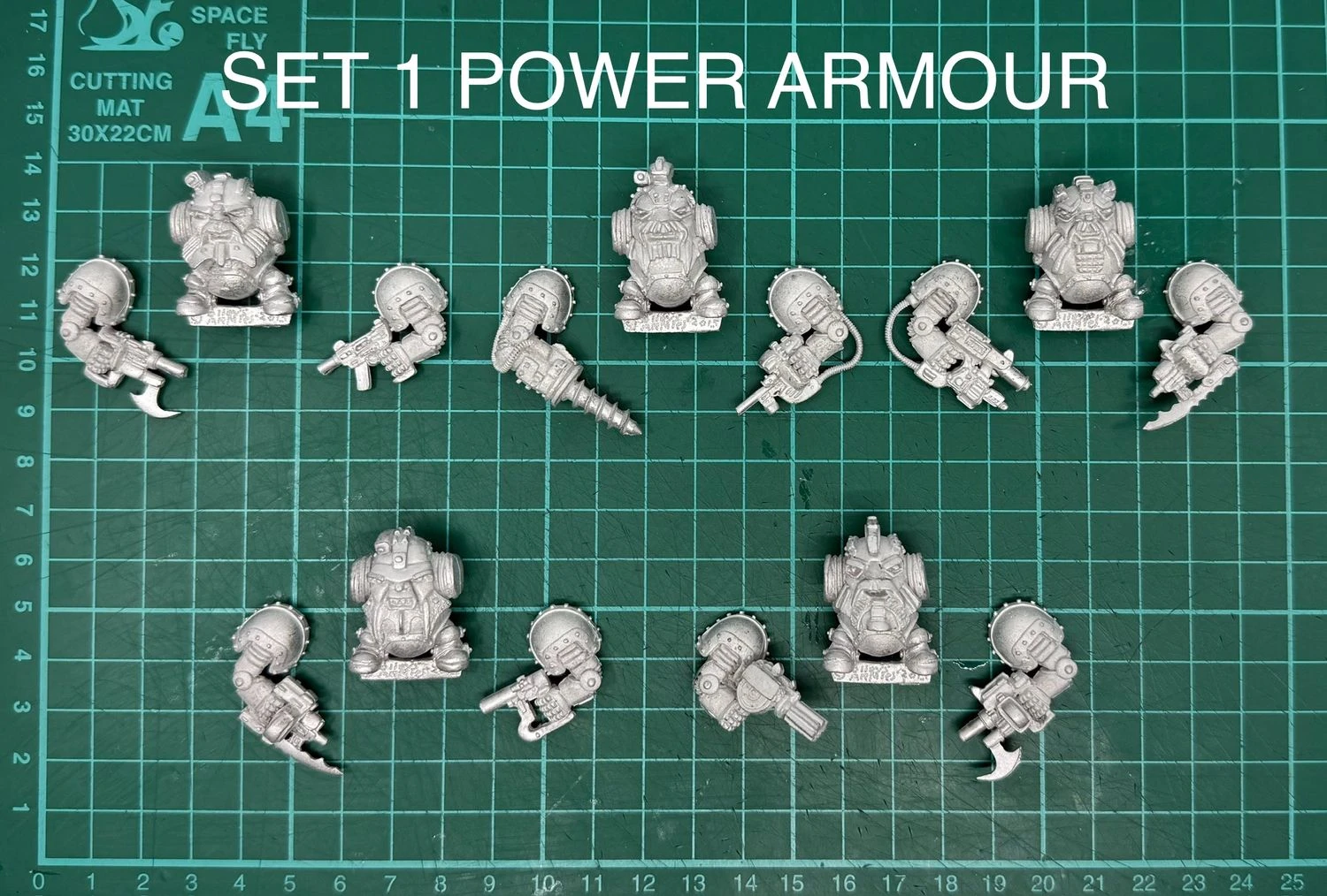 A collection of multipart, white metal miniatures featuring armoured dwarves, arranged on a green cutting mat with a grid pattern. The top row displays various dwarf figures in sci-fi power armour, each equipped with different weapons, including a drill and firearms. The text "SET 1 POWER ARMOUR" is prominently displayed above the miniatures, indicating the theme of this set.