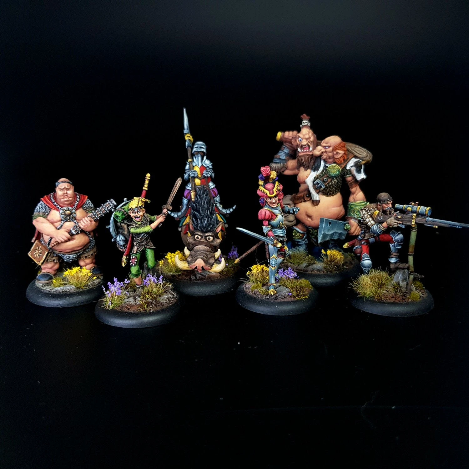 A group of eight intricately painted Moonstone miniatures stands against a black background. The characters include a muscular figure with a weapon, a green-skinned goblin in colorful attire, a knight in armor, and various other fantastical beings, each displaying unique features and vibrant colors. They are positioned on circular bases adorned with tufts of grass and small flowers, showcasing the detailed artistry of the miniatures.