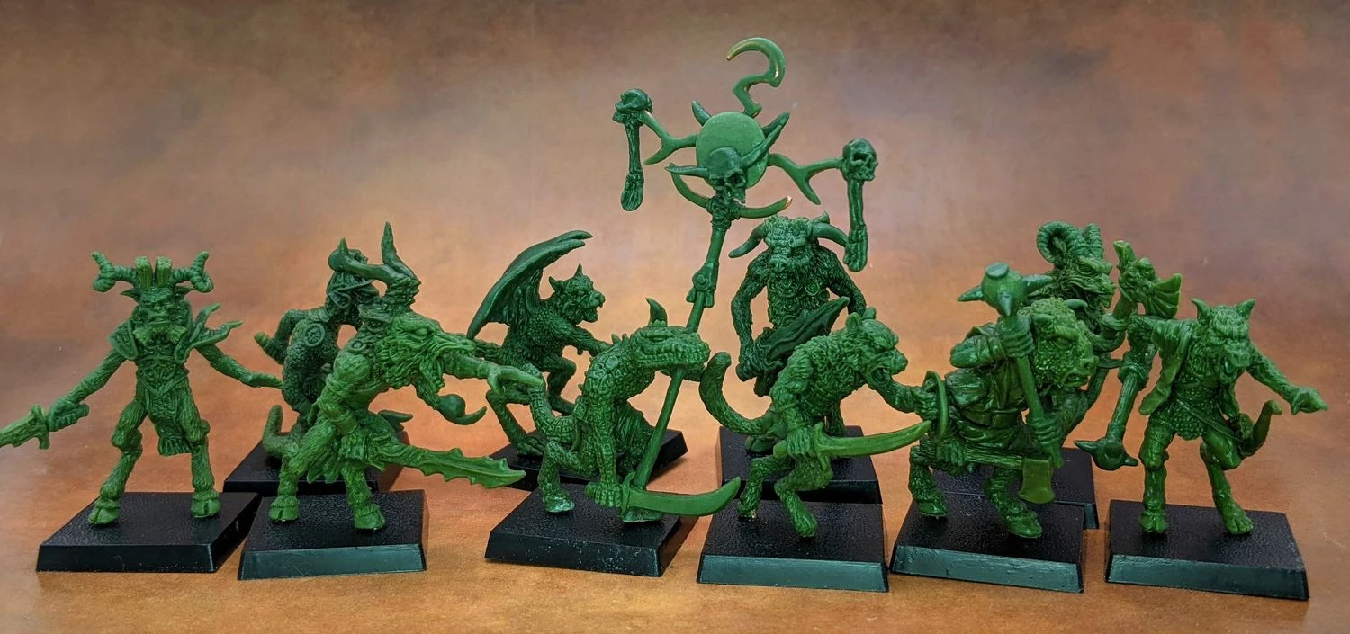A collection of old school miniatures from Satyr Art Studio, featuring various fantastical creatures in a vibrant green color. The figures include beastmen with distinct features such as horns and claws, some wielding weapons, and a central figure holding a staff topped with a skull and a crescent. They are displayed on black bases against a warm, textured background.