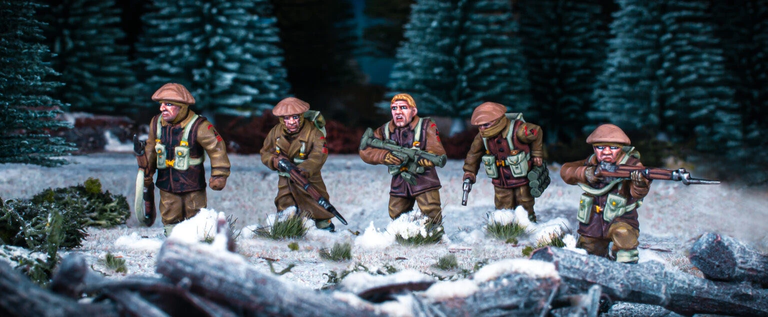 Three British and Canadian winter infantry figures are depicted in a snowy landscape, surrounded by patches of grass and rocks. Each figure is dressed in warm winter gear, with various poses: one holds a rifle, another has a sidearm, and the third is equipped with a machine gun. The background features dark green trees, enhancing the wintery atmosphere of the scene.