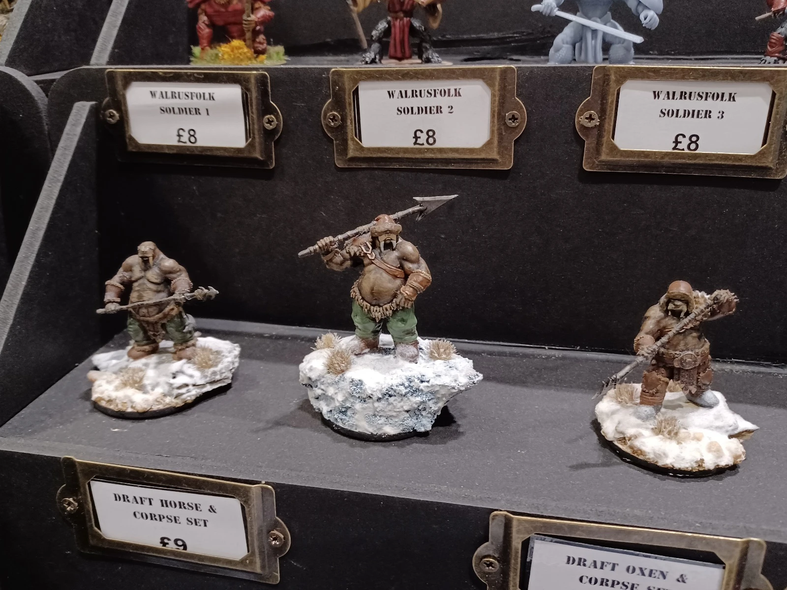 A display of fantasy miniatures features three distinct walrusfolk soldiers, each mounted on snowy bases. The central figure, Walrusfolk Soldier 2, wields a spear and is depicted with a muscular build, wearing green pants and a bare upper body. The other two figures, labeled Walrusfolk Soldier 1 and Walrusfolk Soldier 3, showcase different poses and attire, with one holding a weapon and the other appearing more rugged, all set against a dark backdrop that highlights their details.