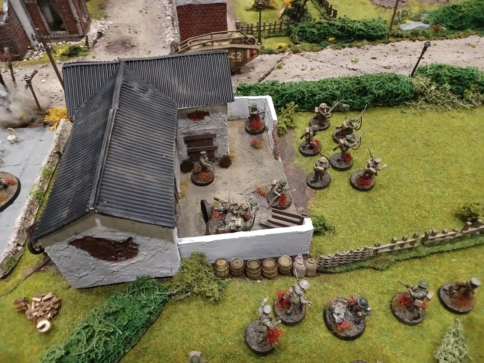 A detailed tabletop wargaming scene depicts a World War I battle setup, featuring a partially destroyed building with a slanted roof and a small group of soldiers positioned around it. In the foreground, several miniature soldiers are engaged in combat, some taking cover behind barrels and others advancing with weapons drawn. The terrain includes grassy areas, scattered foliage, and a dirt path, creating a realistic battlefield atmosphere.