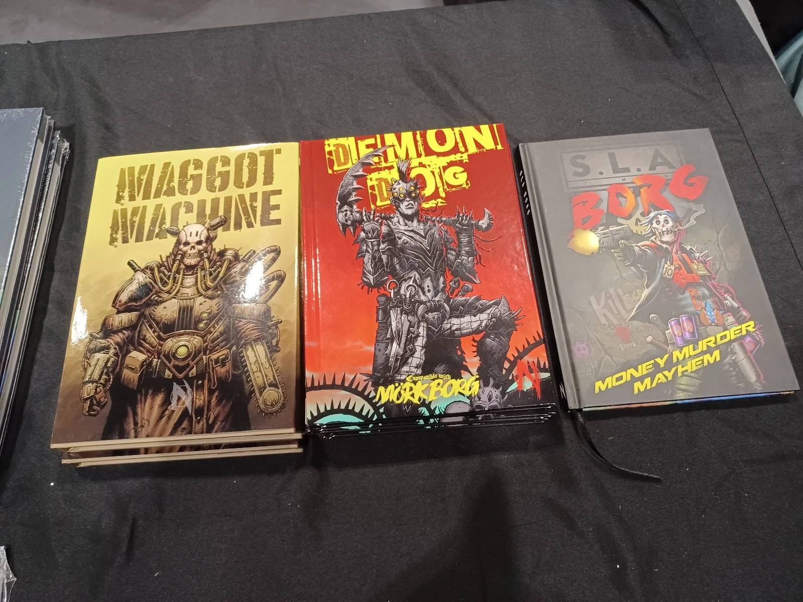 Three tabletop role-playing game books are displayed on a black table. From left to right, the first book titled