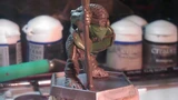 A small, detailed miniature figure stands on a square base, holding a staff and wearing a green mask. The figure is dressed in textured armor, showcasing intricate details like scales and straps. In the background, various paint pots and tools are visible, indicating a workspace for painting or modeling miniatures.